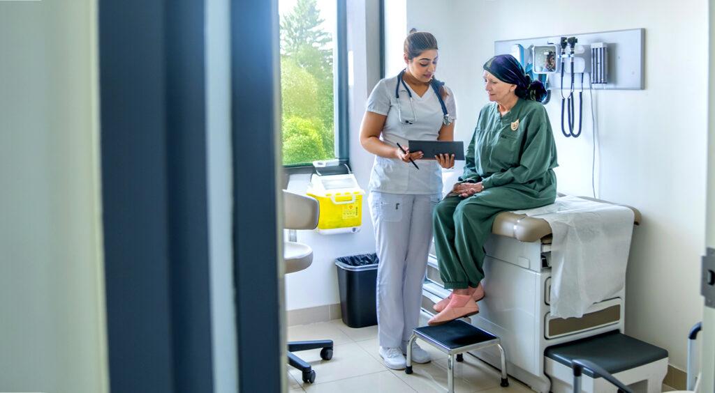 A senior woman who is battling cancer, sits up on an exam table during an appointment. She is dressed comfortably and wearing a headscarf to keep her warm as she talks with the doctor.