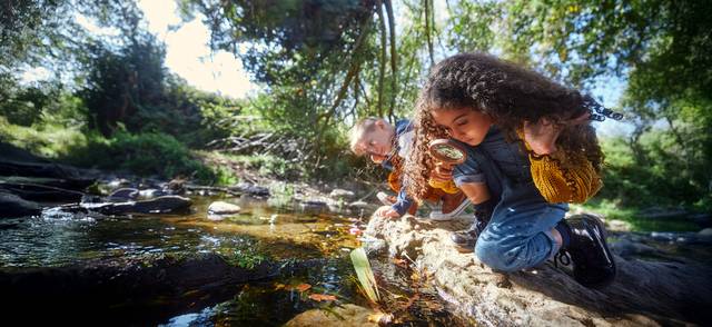 Two young people crouch down and look at river with magnifying glass.