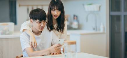 A photo of a young couple at a kitchen table looking at a phone with a surprised look on their faces