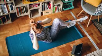 Woman in striped shirt and tights exercising on yoga mat while looking at tablet.