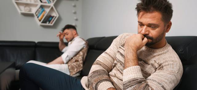 Two men sitting apart on a couch, one in a beige sweater deep in thought, the other in a white shirt resting head in hand.