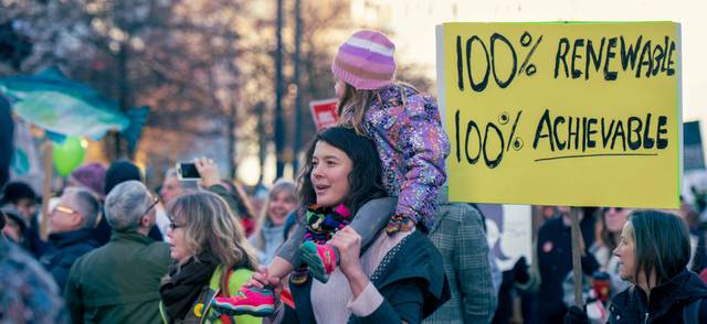 A group of people at a climate change protest. In the centre of the photo is a young woman with a child on her shoulders