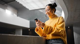 An Asian woman is smiling as she texts on her smartphone while reading on it.