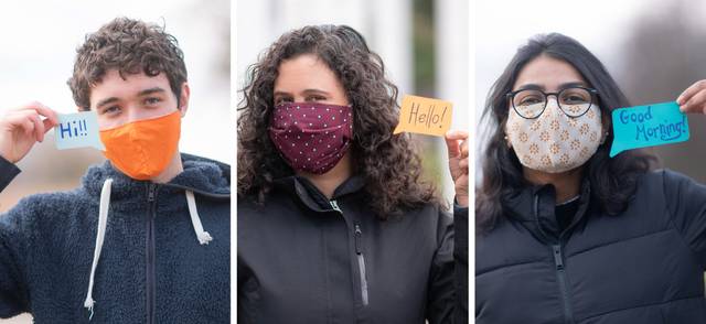Three people wearing COVID-19 face masks say hello using signs