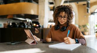Young adult woman taking notes while using a tablet in a modern cafe setting.