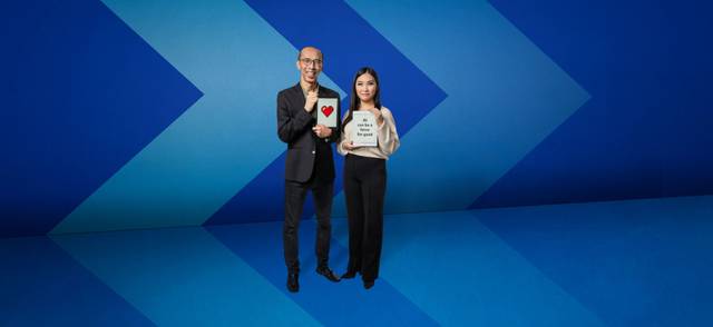 Two people stand smiling in front of a blue chevron background. Dr. Raymond Ng holds a digital printout of a heart, and Christy Kwok holds a sign that reads &quot;AI can be a force for good&quot;.