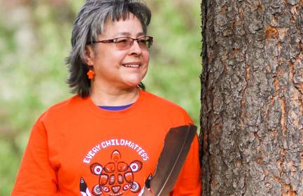 Phyllis Webstad, founder of Orange Shirt Day, stands next to a tree and holds a feather.