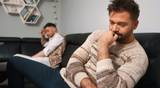 Two men sitting apart on a couch, one in a beige sweater deep in thought, the other in a white shirt resting his head in his hand.