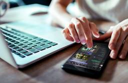 A person taps a phone next to an open laptop.