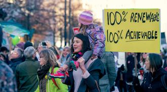 A group of people at a climate change protest. In the centre of the photo is a young woman with a child on her shoulders