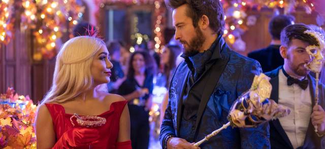 Woman in red dress holding cocktail and man in blue suit holding a mask at masquerade party surrounded by lights