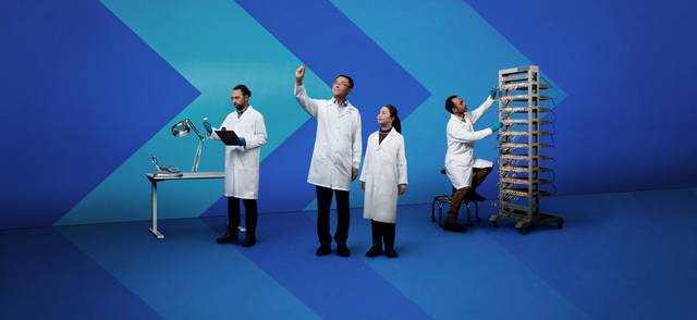 Four people in lab coats work with batteries in a stylized lab with chevron backdrops.