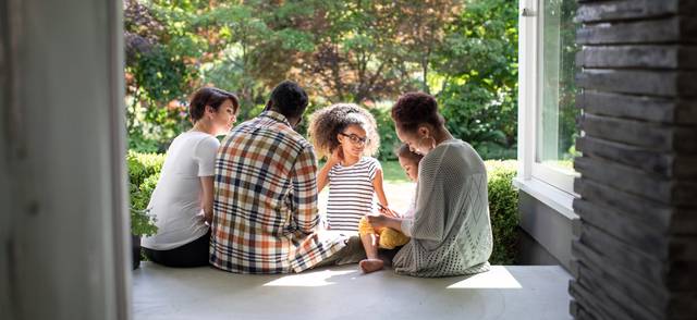 A Black family with a mother, father and three children sit beside one another on the porch.