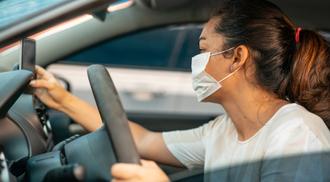 A woman wearing a mask in the driver's seat looking at a cell phone.