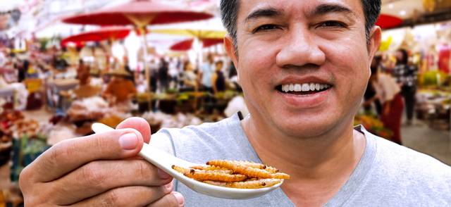 A person grins and holds a spoon full of small bamboo caterpillars at a market in Thailand.