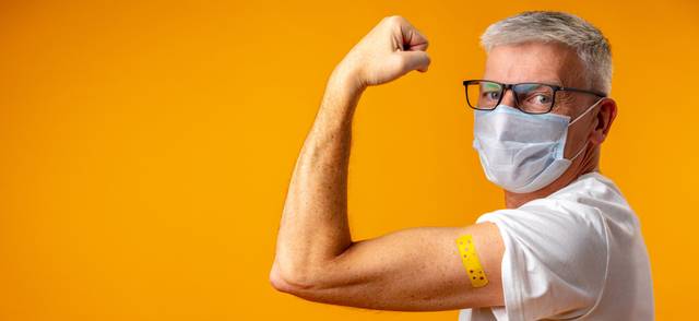 A person posing with their arm flexed with a band-aid over an injection against orange background.
