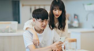 A photo of a young couple at a kitchen table looking at a phone with a surprised look on their faces
