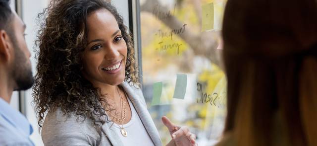Young black woman holding a conversation with peers next to a window with ideas posted on notes