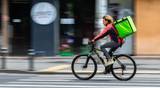 A cyclist wearing a red and black jacket rides quickly down a city street, carrying a large green delivery backpack, with blurred buildings in the background.